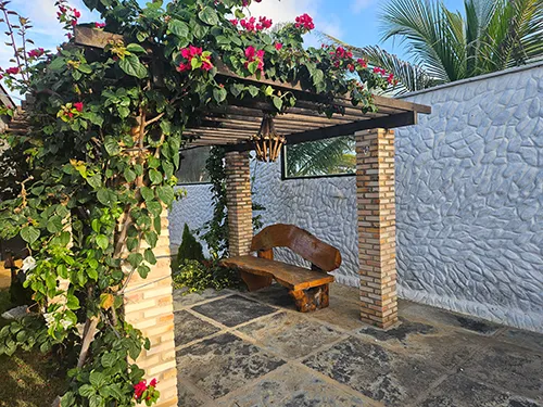 Recanto Charmoso com Pergolado - Pousada Bellíssima Itália Recanto com banco de madeira sob pergolado coberto por flores nos jardins da Pousada Bellíssima Itália, Luis Correia.