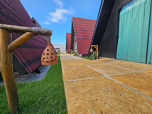 Pousada Bellissima Italia: Charme e Arquitetura Única dos Nossos Chalés Perspectiva do corredor externo entre os chalés triangulares da Pousada Bellissima Italia, mostrando o design arquitetônico e o paisagismo sob um céu azul com nuvens.